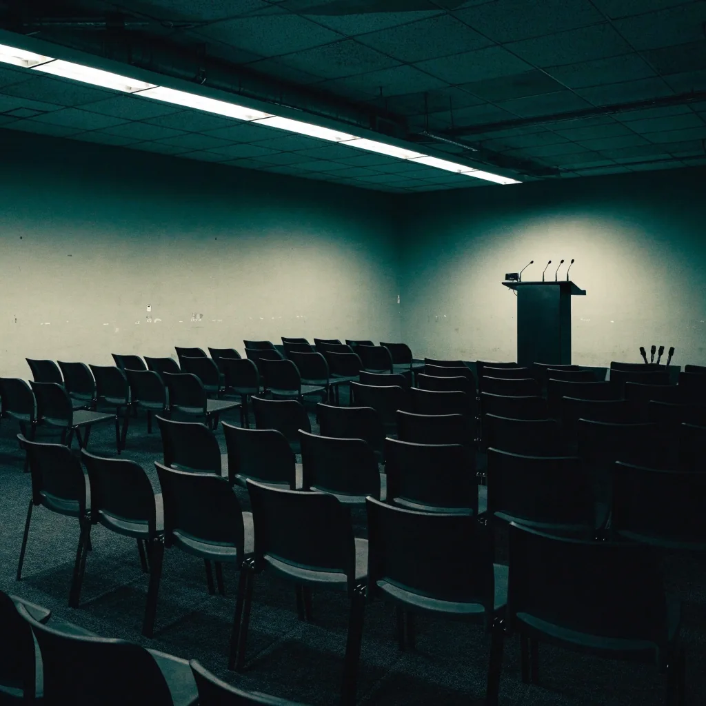 A dim press briefing room from the back of the hall, empty seats facing a lectern with microphones