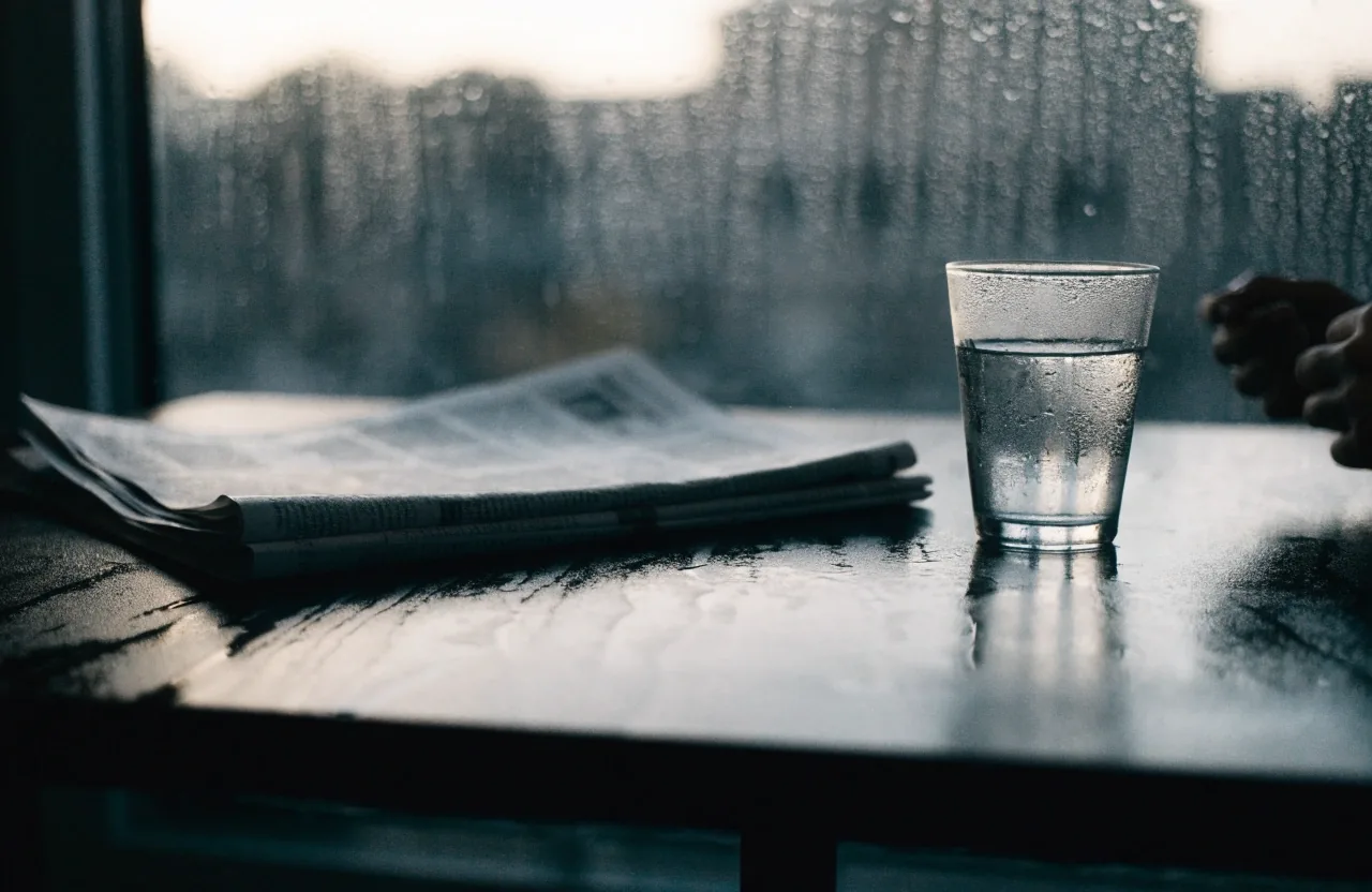 Folded newspapers on a wet cafe table beside a glass of water in morning light